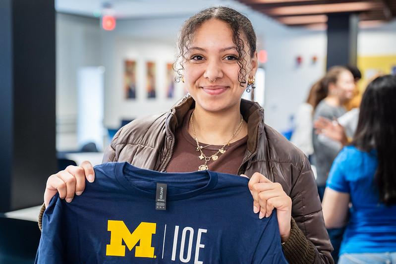  Female student with curly black hair pulled back, dressed in a brown jacket holding a navy blue t-shirt with M-IOE printed on it in maize and white. In the background there is a blurred out classroom and students talking. 