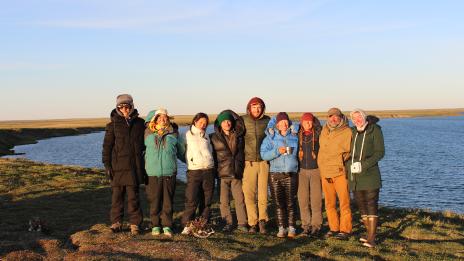 Ten members of SEAS in insulated jackets and boots pose arm-in-arm on grassy ground next to a calm, winding river, with expansive plains and a blue sky behind them.