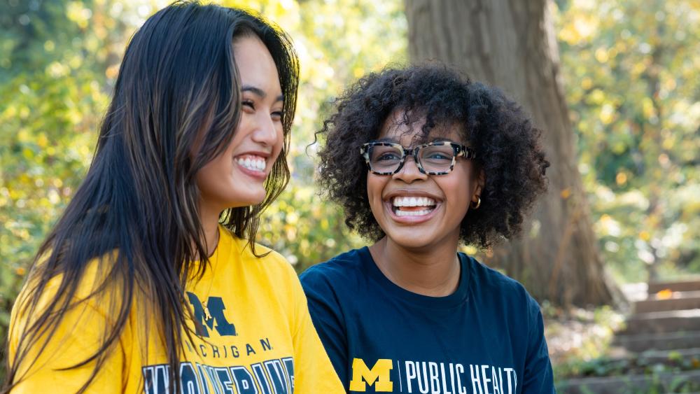  A photograph of two students sitting on steps in a wooden area. One of the students has a University of Michigan yellow Wolverines t-shirt on and the other student has a navy blue Michigan Public Health t-shirt on and they are both smiling and laughing. 