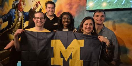 Five people holding a University of Michigan Alumni Association banner in front of a colorful mural with a football game on a nearby TV.