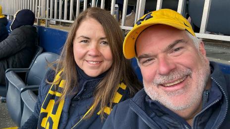 Maureen and Jim Lico sit together in stadium seats, wearing University of Michigan colors. 