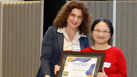 Two people pose together holding a framed University of Michigan award certificate.