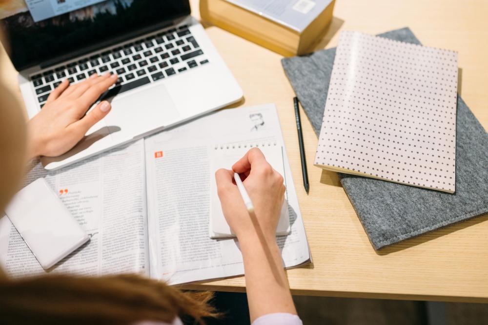 Person studying at a desk, typing on a laptop while taking notes beside an open book, notebooks, and school supplies. 
