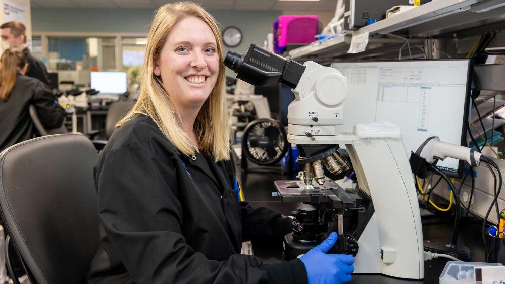 A smiling researcher wearing gloves sits at a microscope in a laboratory workspace. 