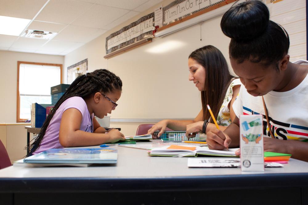 A female teacher sits at a table with two female students. She is working with one student in the background while the student in the foreground writes in a notebook. 