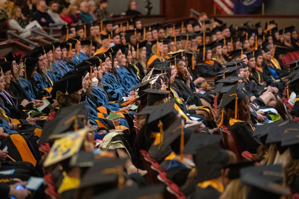 Crowd of nursing students sittin in caps and gowns at graduation listening to speaker.