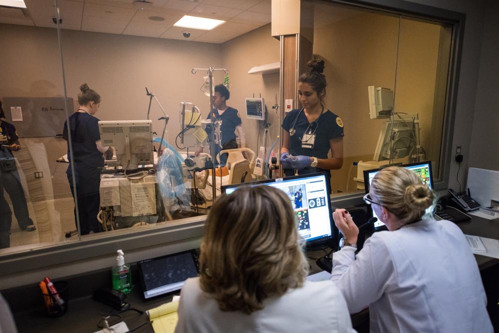 Nursing instructors observe nursing students in simulated hospital room lab.