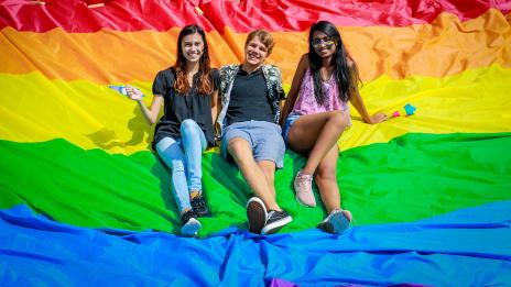 Three students pose outside on a rainbow flag