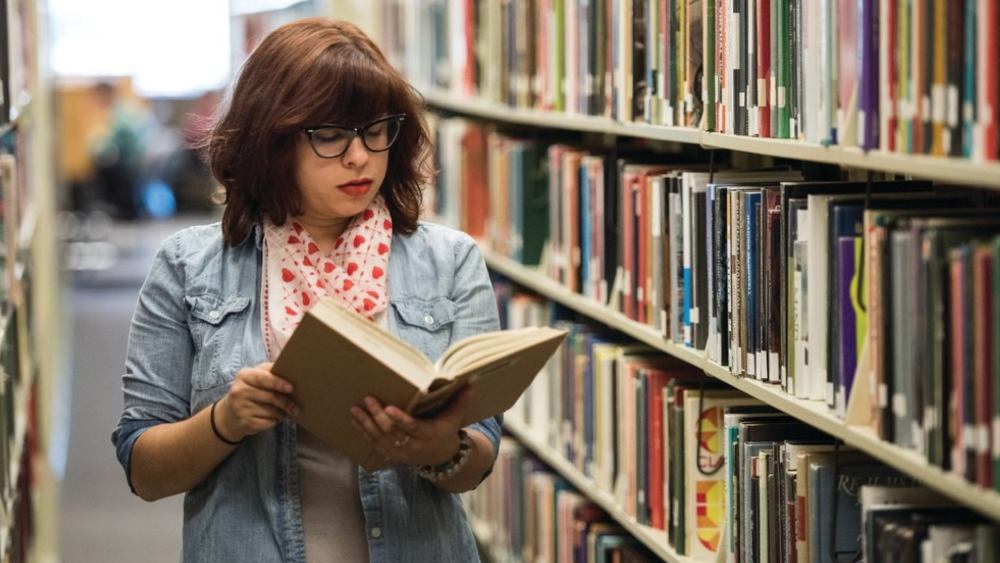 Student in standing in library aisle reading a book