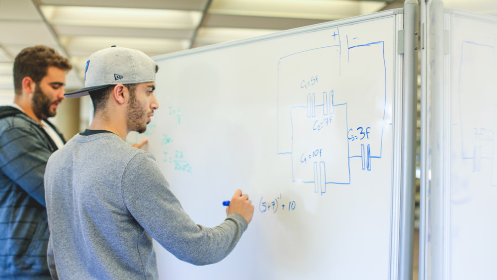 Two students standing in front of a whiteboard solving a math problem