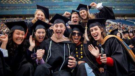 Eight graduates in black caps and gowns cheer and raise their fists together at a crowded stadium commencement ceremony.