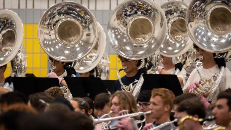 Musicians perform in a crowded indoor ensemble, with a row of reflective silver sousaphones towering above trumpet players in the foreground.