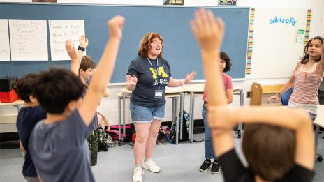 A teaching intern smiles and gestures while leading an energetic classroom activity as students gather around with raised hands.