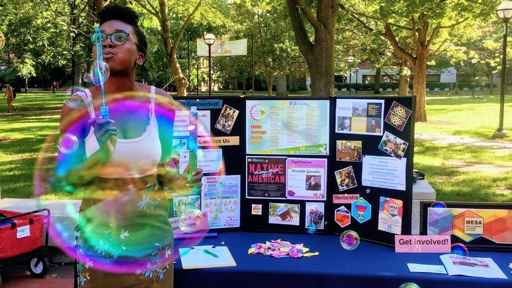 A student blows a bubble in front of a table for the Multi Ethnic Student Affairs office 