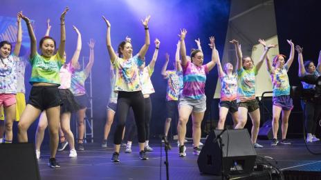 A group of females on a stage with their hands raised, singing and dancing