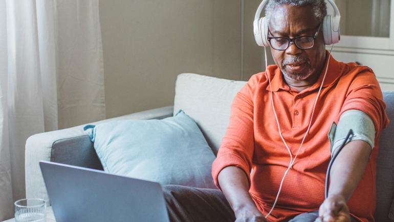 An older male, wearing headphones and sitting in front of an open laptop, takes his blood pressure