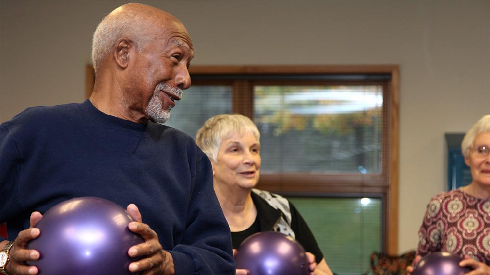 An older male, holding an exercise ball, talks to two older females in the background, who are also holding exercise balls