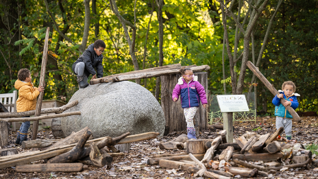 Group of kids playing in children's garden, climbing on boulders, and holding large sticks. 