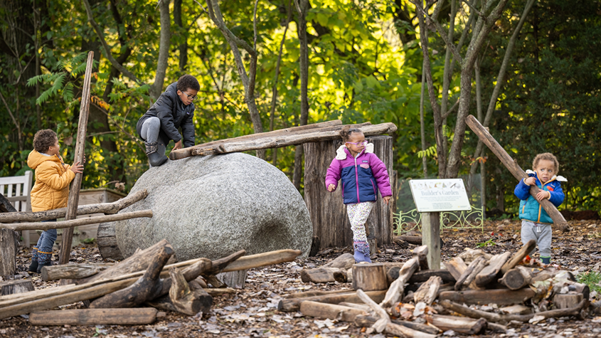 Group of kids playing in children's garden, climbing on boulders, and holding large sticks. 