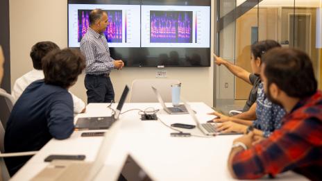 A man stands at the front of a room near a digital display screen. Several people are looking toward him. One of the people is pointing at the screen