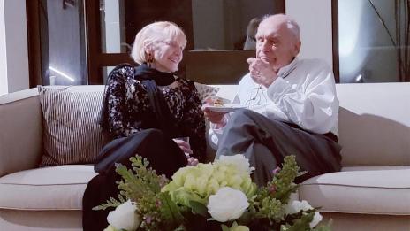 Pris Rogers and Gunter Dufey, dressed in formal cocktail party clothing, sit together on a sofa, engaged in conversation with one another.