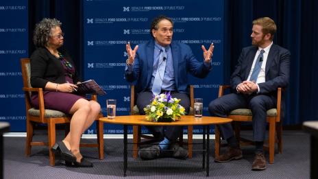 A woman and two men sitting in chairs around a coffee table having a discussion in front of a backdrop that reads “Gerald R. Ford School of Public Policy.”