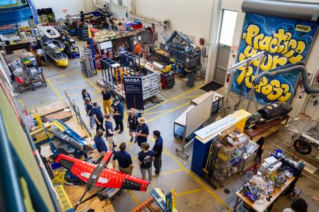 Overhead shot of 12 students and faculty wearing blue shirts in a garage surrounded by tools and vehicle prototypes
