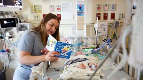 A nurse reads a book to an infant laying in a hospital crib at C.S. Mott Children’s Hospital.