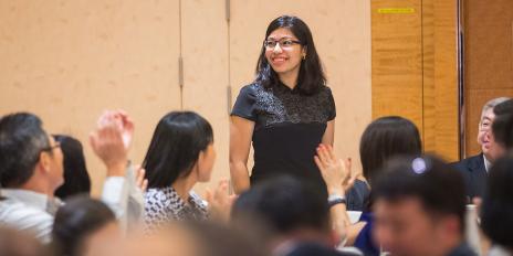 A young woman stands in a room full of people. Some of them are clapping for her.