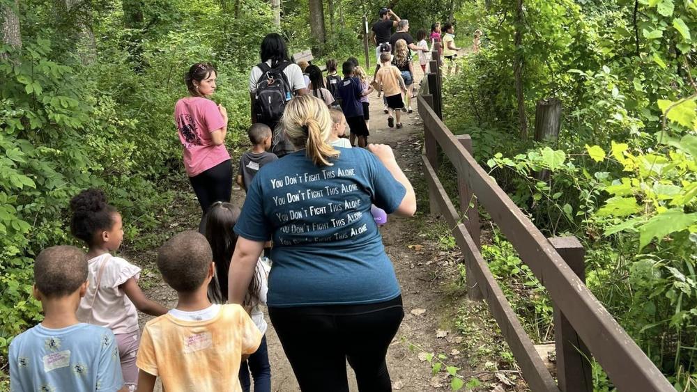 A group of children and a few adults walking away from the camera on a trail in the forest. 