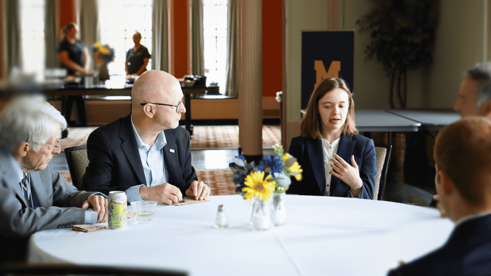 A female studen in a navy blazer sits at a round table speaking to four older men who are looking at her.